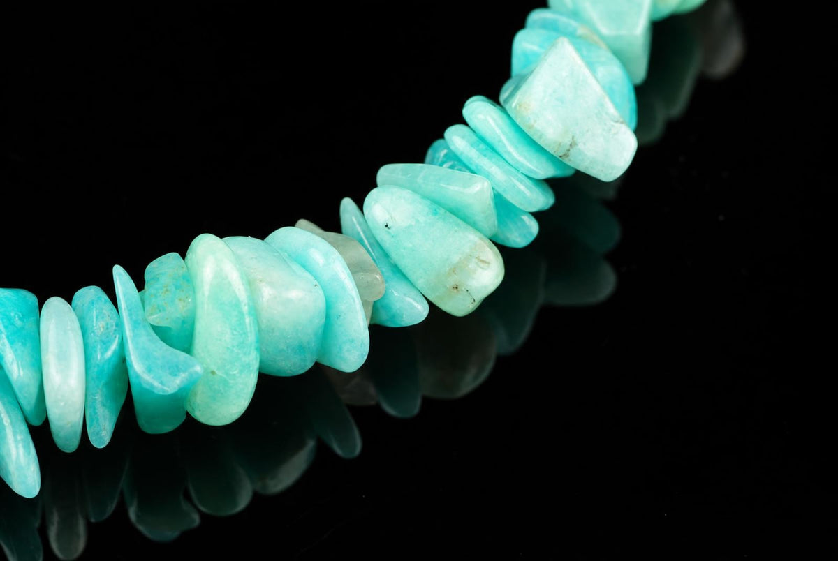 A bracelet made of polished, irregular chips of light blue-green amazonite stone arranged in a line on a dark reflective surface.