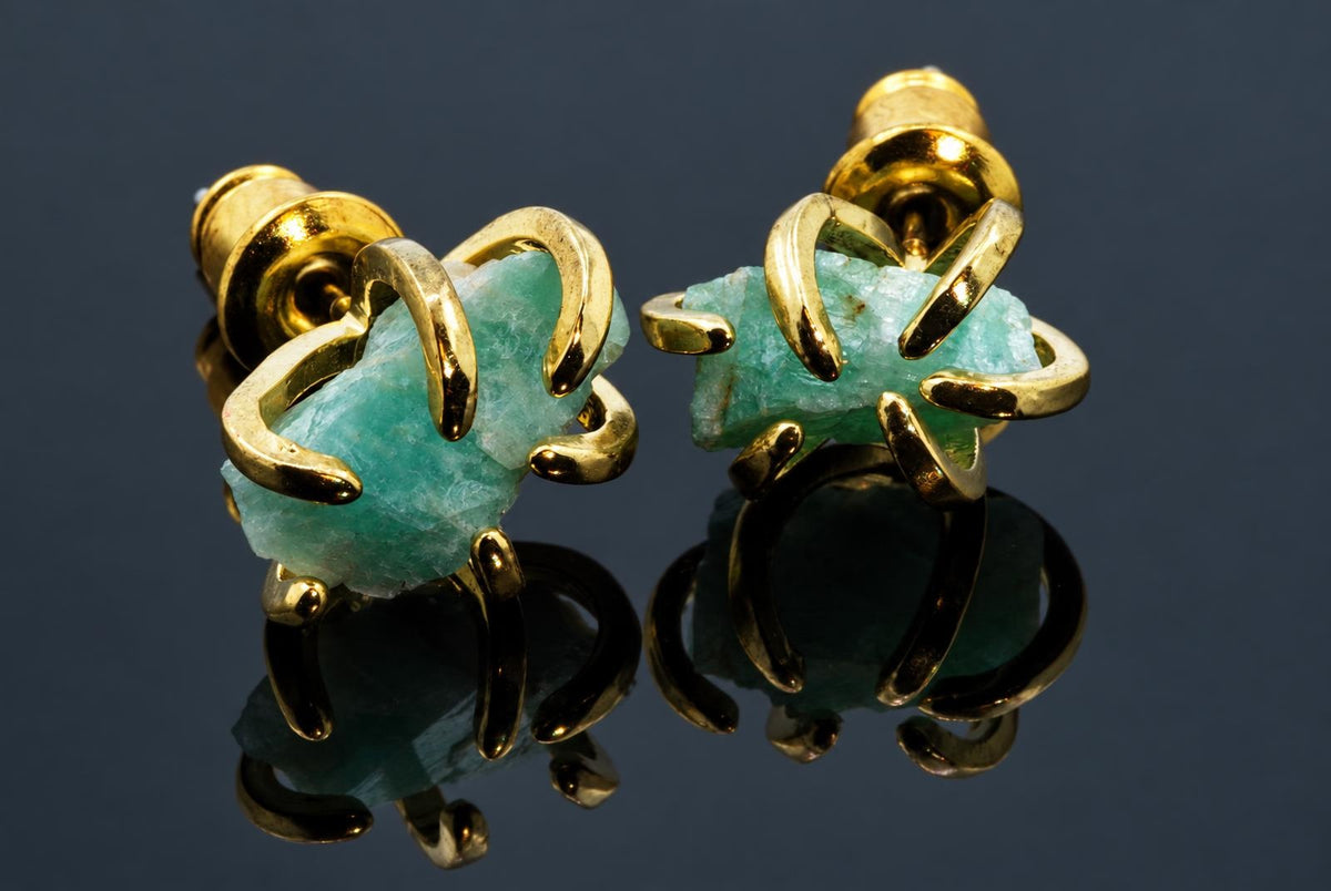 A pair of gold-toned stud earrings with curved metal prongs holding rough, light green amazonite stones on a dark reflective surface.