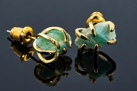 A pair of gold-toned stud earrings with raw, pale green amazonite stones held by curved metal claws on a dark reflective surface.