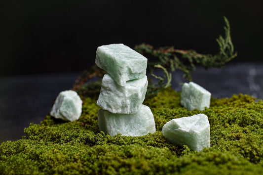 Stacked and scattered pale green Amazonite India raw stones on a mossy surface with a dark background.