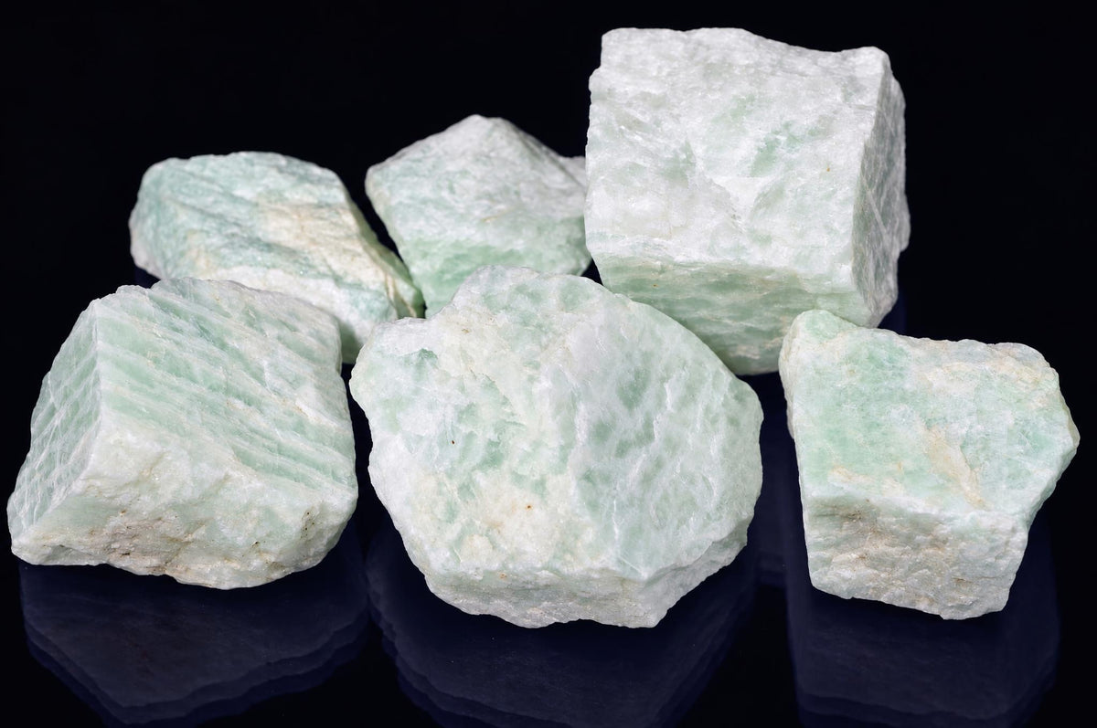 Six raw Amazonite India mineral specimens with pale green and white crystalline textures on a dark reflective surface.