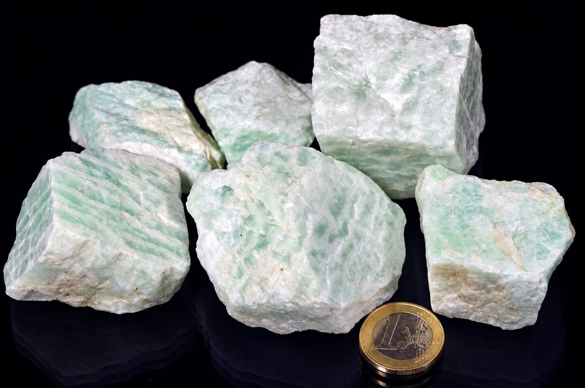 Several rough pale green amazonite pieces show white streaks and angular broken surfaces, arranged beside a coin on a reflective black background.