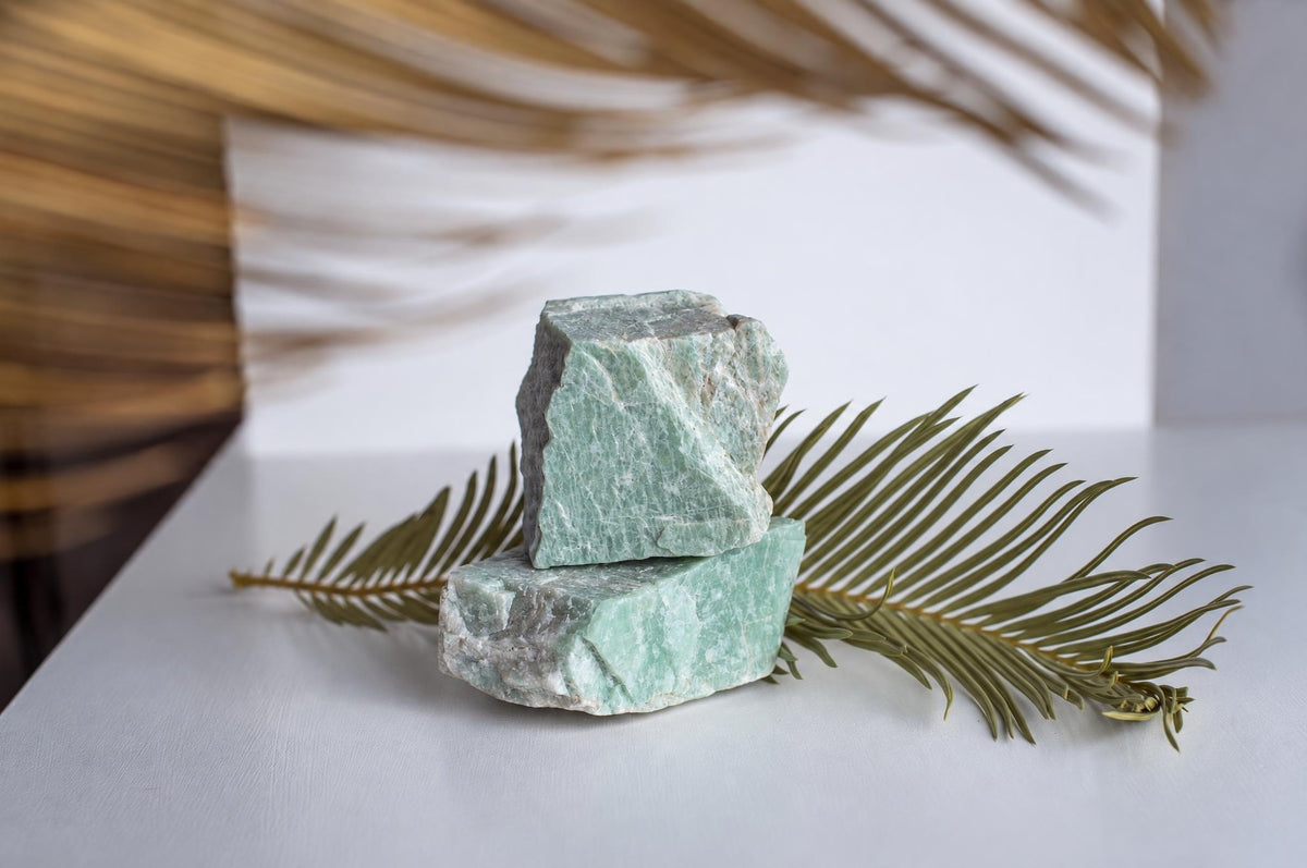 Two rough, light green Amazonite stones stacked on a white surface with a dried palm frond nearby.
