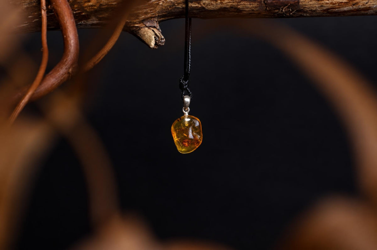 A polished amber pendant with a silver bail hangs from a black cord against a dark background, suspended near a wooden branch.