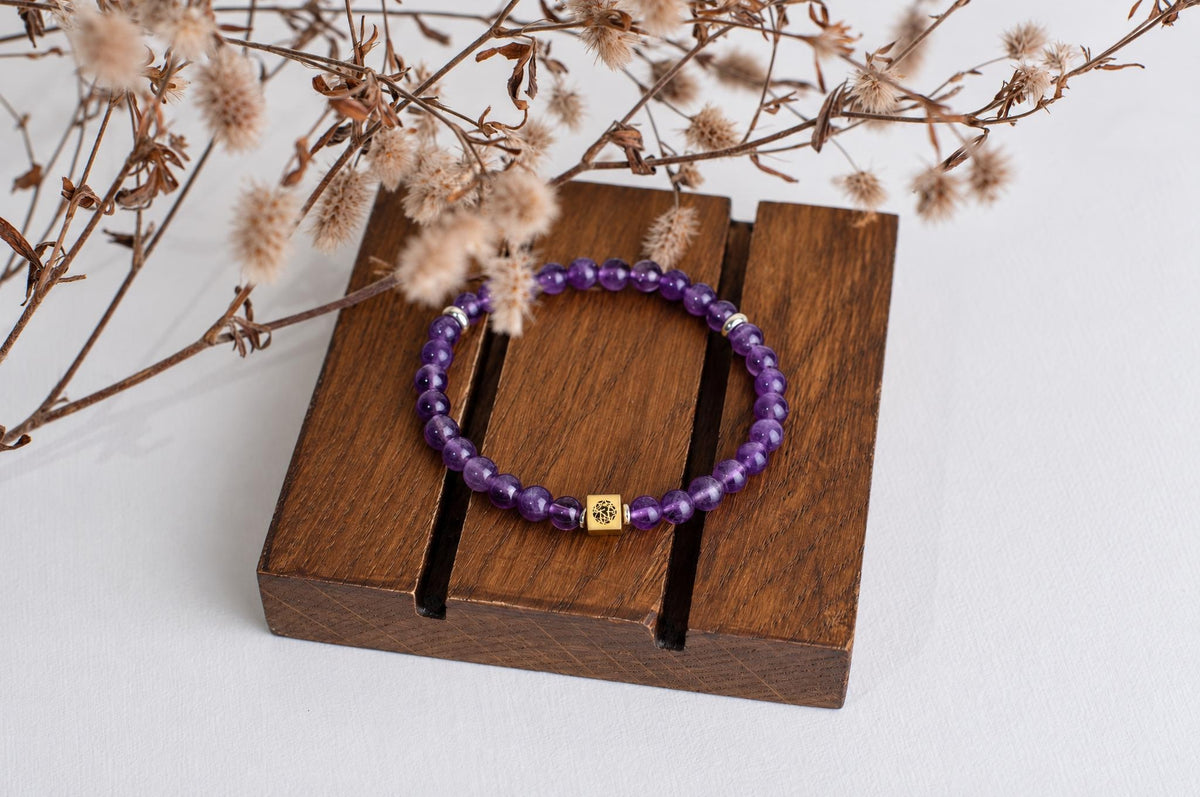 A bracelet with round purple beads and a gold-toned square charm rests on a dark wooden surface, partially covered by dried flowers.