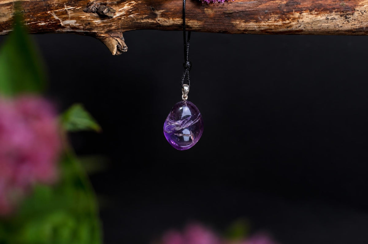 A purple, teardrop-shaped pendant with a silver-toned bail hangs from a black cord on a wooden branch against a dark background.