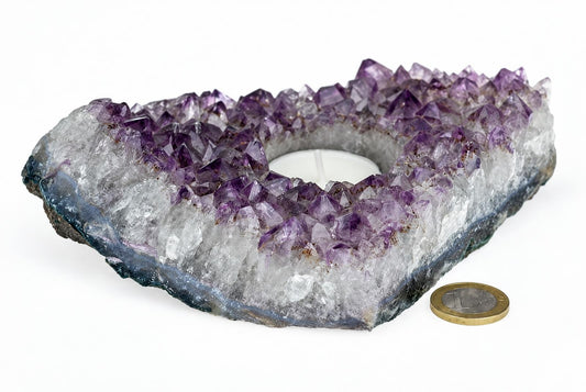A heart-shaped amethyst geode candle holder with purple and clear crystals, a white tea light inside, on a pale surface next to a coin.
