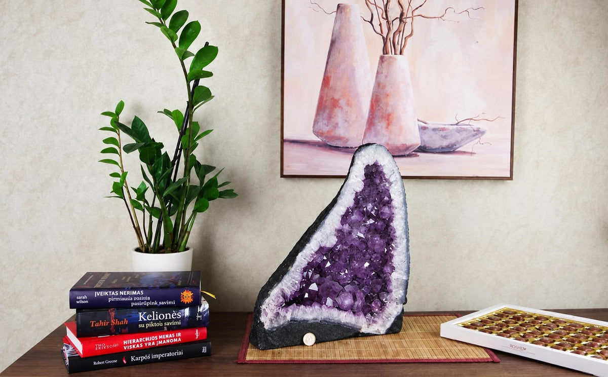 A large amethyst geode slice with purple crystals and white rim sits on a bamboo mat beside books, a plant, and chocolates.