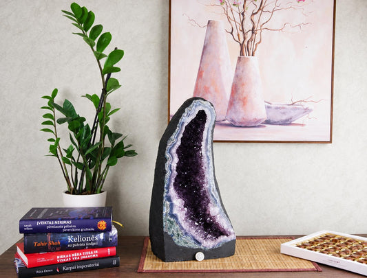 A large, dark geode with purple and white crystals sits on a wooden surface next to books and a plant.