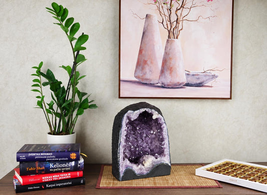 A large amethyst geode with purple crystals sits on a dark surface, next to books and a plant.