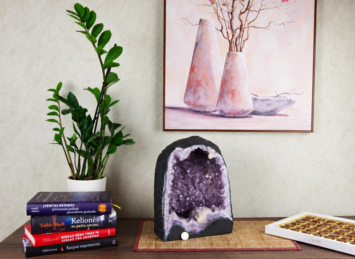 A large amethyst geode with purple crystals sits on a dark surface next to books and a plant.