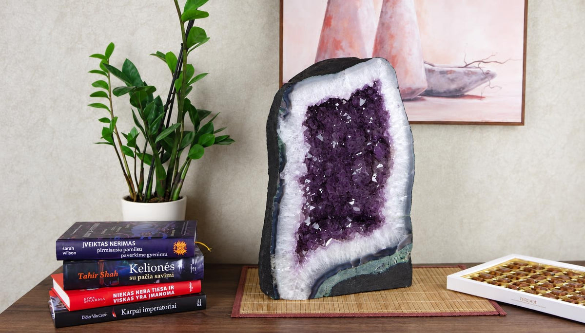 A tall amethyst geode with purple crystals and a pale rim stands beside books and a leafy plant.