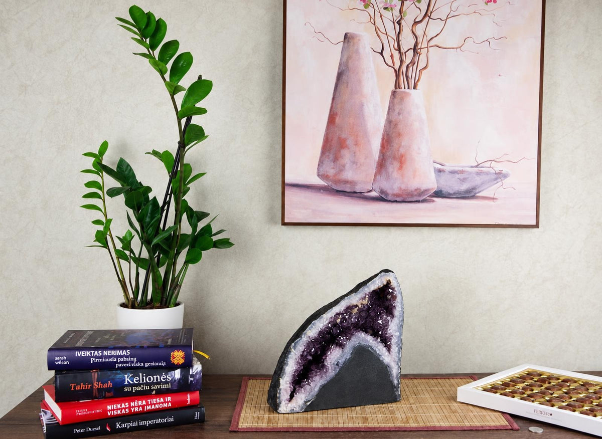 A large amethyst geode slice with purple crystals sits on a bamboo mat beside books and chocolates.