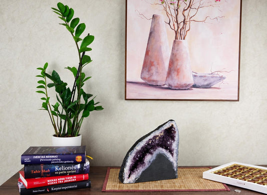 A large amethyst geode slice with purple crystals sits on a bamboo mat beside books and chocolates.