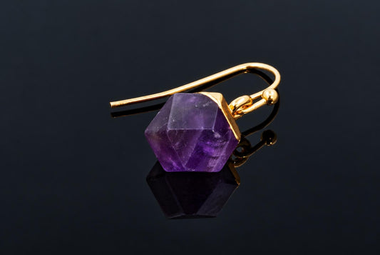 A gold-coloured hook earring with a faceted purple amethyst cube pendant on a dark reflective surface.