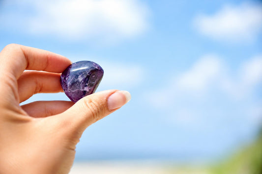 A hand holds a polished, heart-shaped purple amethyst stone against a blurred blue sky and green background.