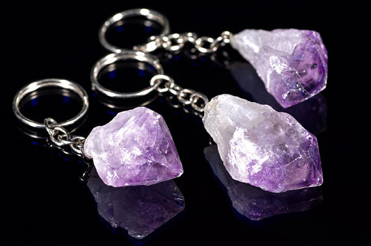 Three silver-toned keychains with rough, purple and white amethyst crystals on a dark reflective surface.