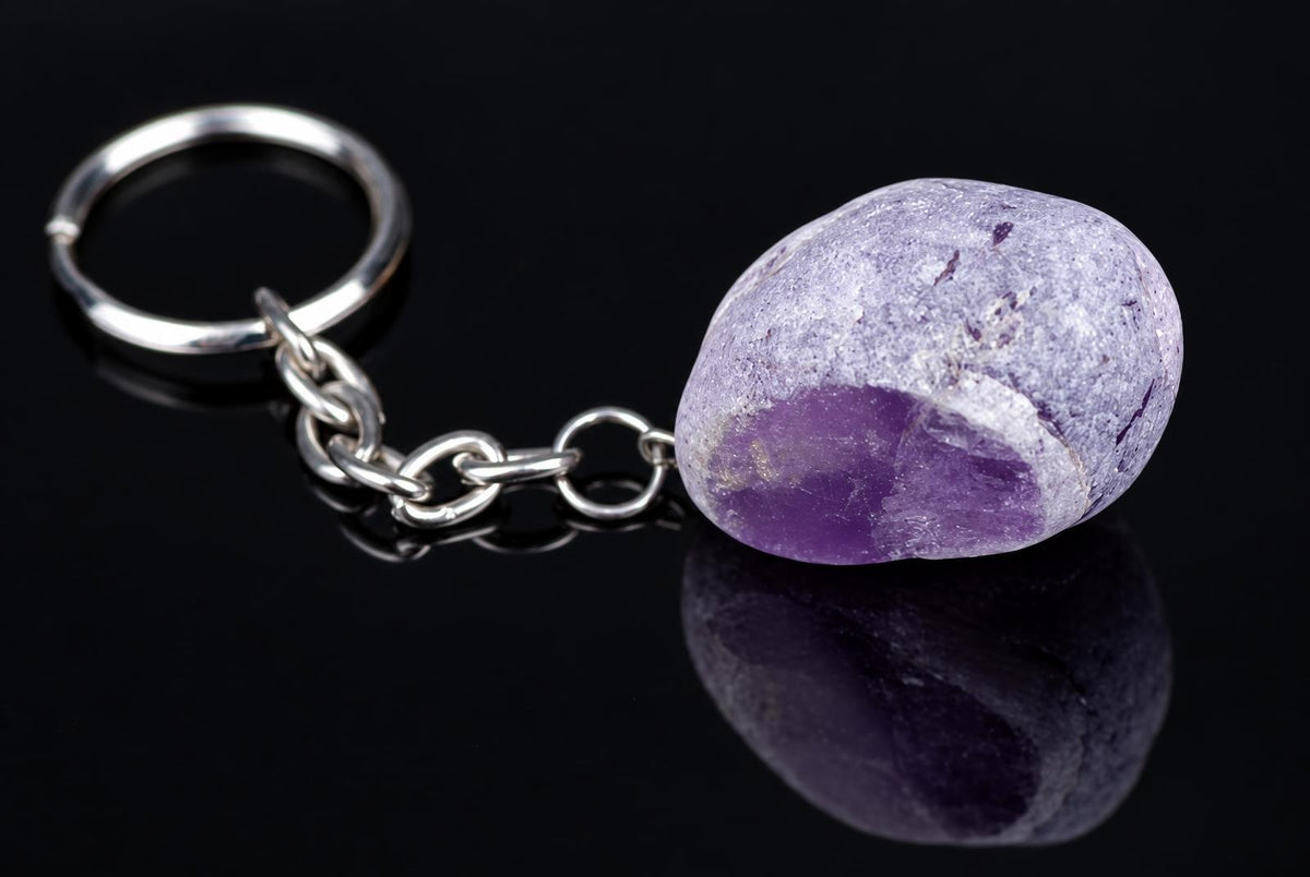 A silver-toned keychain with a chain and ring holds a rough, purple amethyst stone on a dark reflective surface.