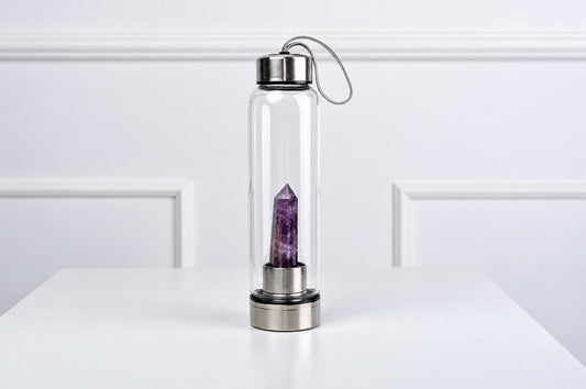 A clear glass water bottle with a silver-toned lid and base, containing a purple amethyst crystal point inside.