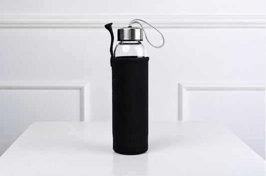 A clear glass water bottle with a silver-toned lid and black fabric sleeve stands on a pale surface.