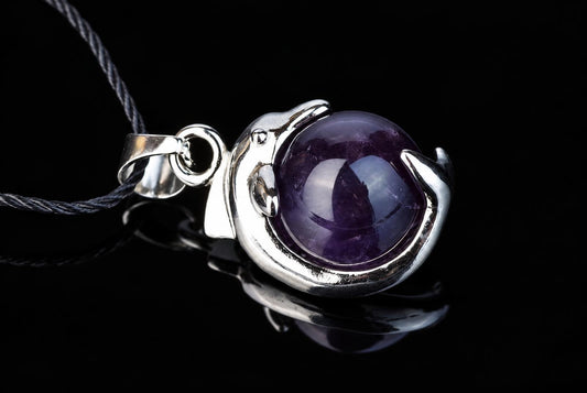 A silver-toned dolphin pendant holds a round, polished purple amethyst stone on a dark cord against a black reflective surface.
