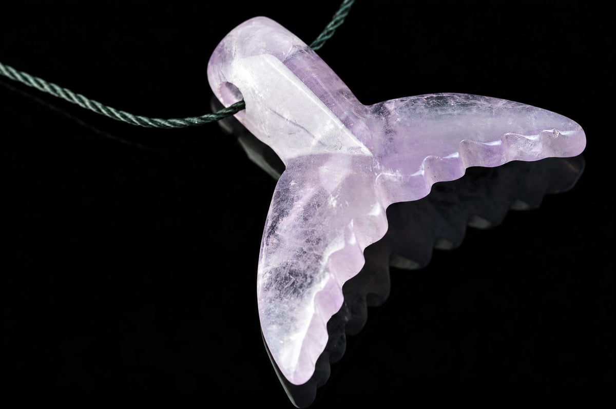 A translucent purple and white carved pendant shaped like a dolphin tail, hanging from a dark green cord against a black reflective surface.