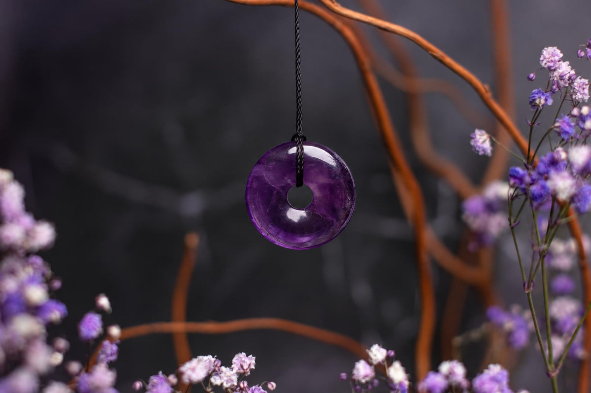 A purple donut-shaped pendant hangs from a black cord, surrounded by small flowers and brown branches.