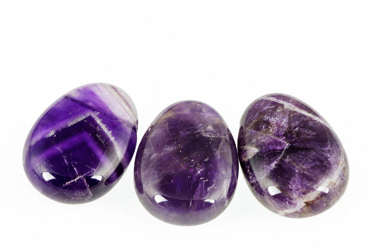 Three polished amethyst stones with deep purple and white banding, arranged horizontally on a pale surface.