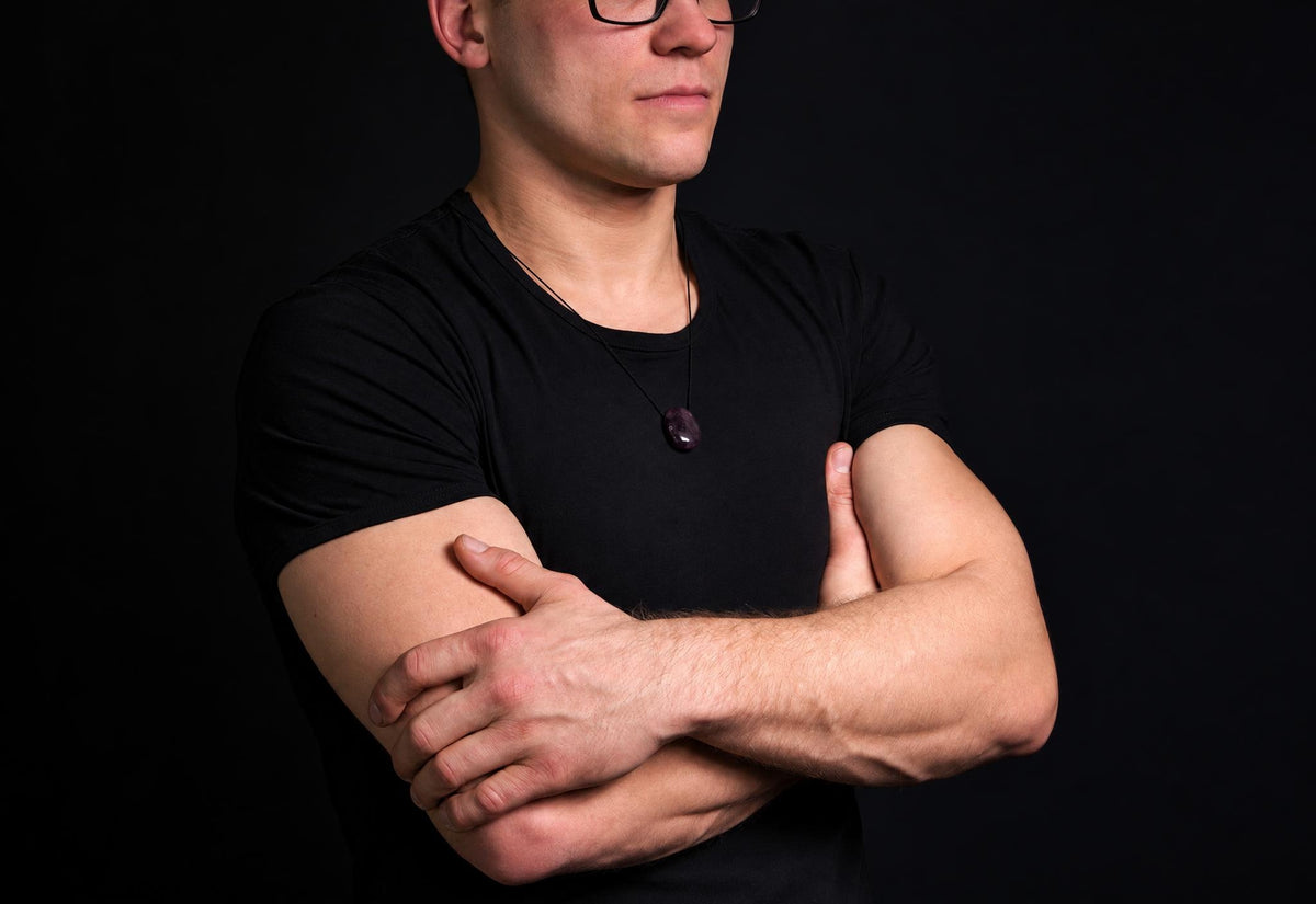 A man in a black t-shirt wears a necklace with a dark purple, oval-shaped pendant against a dark background.