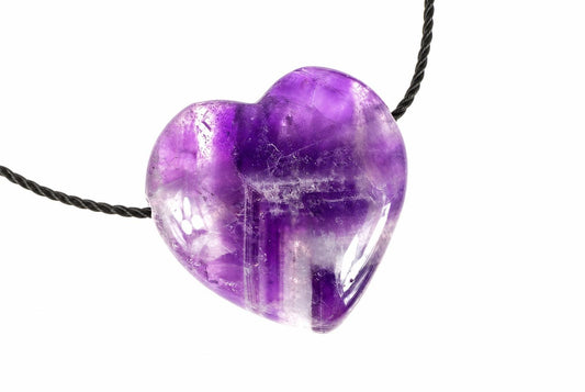 A polished heart-shaped amethyst pendant with purple and clear tones hangs from a dark cord against a white background.
