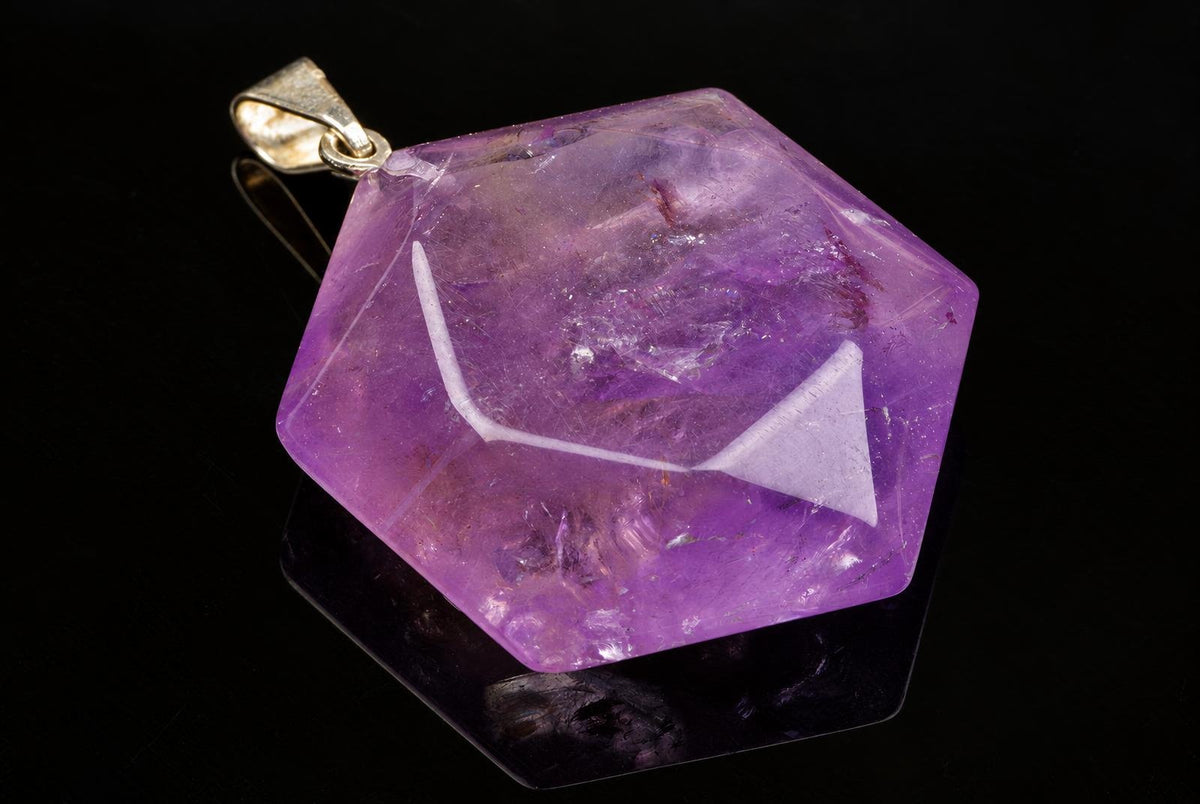 A hexagonal purple amethyst pendant with a gold-toned bail rests on a dark reflective surface.