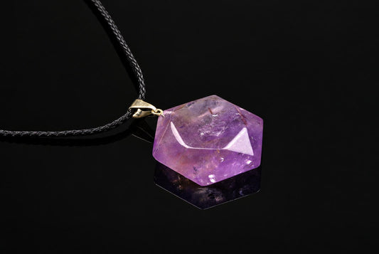 A hexagonal purple amethyst pendant with a gold-coloured bail hangs from a black cord against a dark surface.