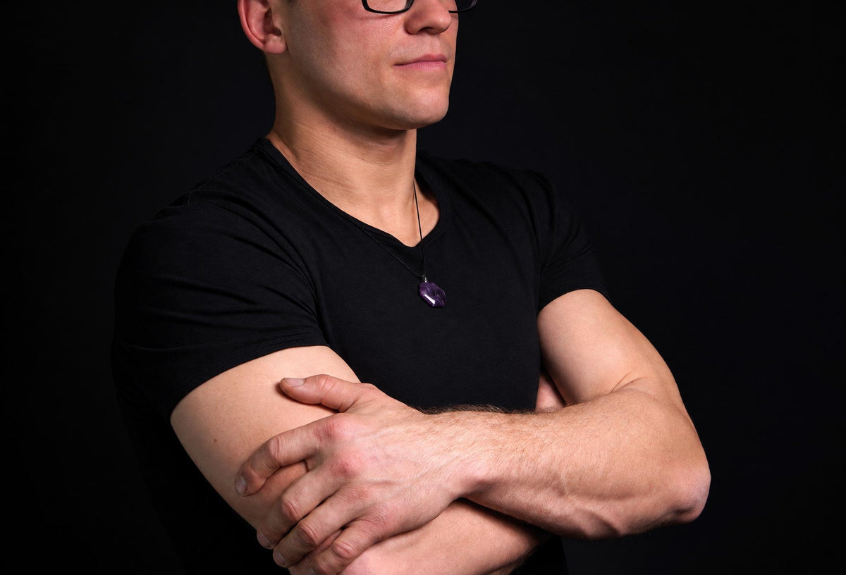 A man in a black t-shirt wears a necklace with a purple, hexagonal pendant against a dark background.