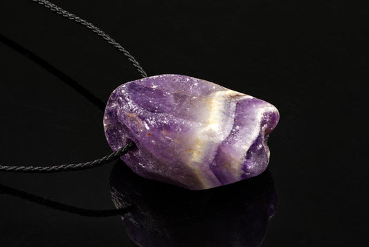 A natural amethyst pendant with purple and white bands on a dark cord, resting on a black reflective surface.