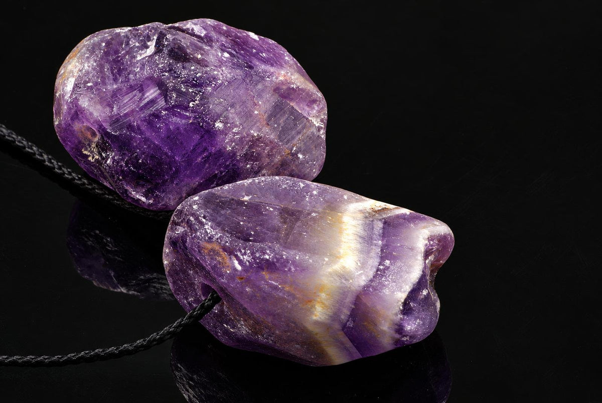 Two rough amethyst stones on a black cord, resting on a dark reflective surface.
