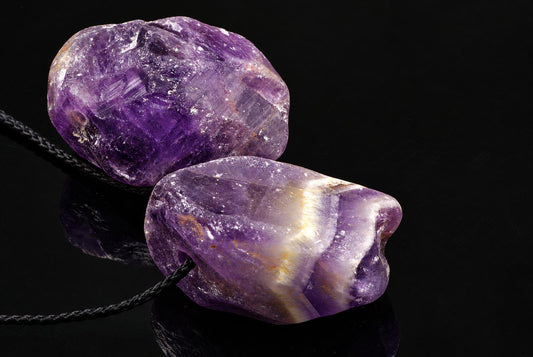 Two rough amethyst stones on a black cord, resting on a dark reflective surface.