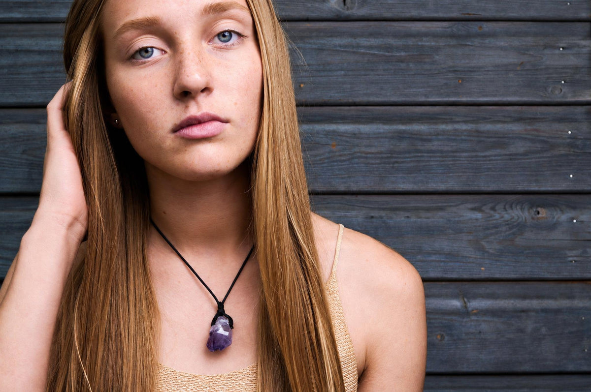 A woman with long blonde hair wears a black cord necklace with a purple amethyst pendant against a dark wooden background.