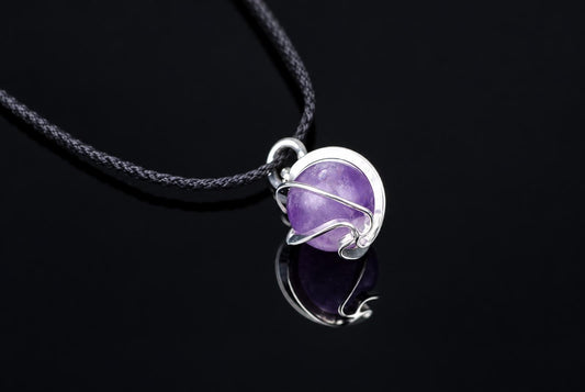 A silver-toned pendant with a round purple sphere, wrapped in wire, on a dark cord against a black reflective surface.