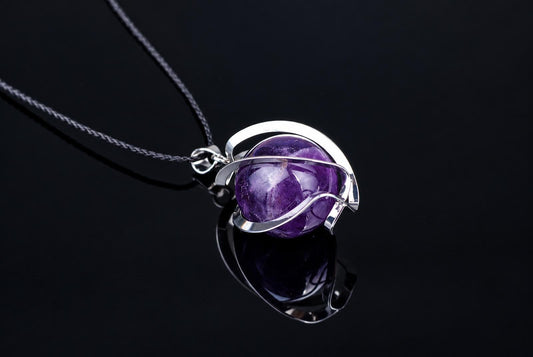 A silver-toned wire pendant holds a round, purple amethyst sphere on a dark cord against a black reflective surface.