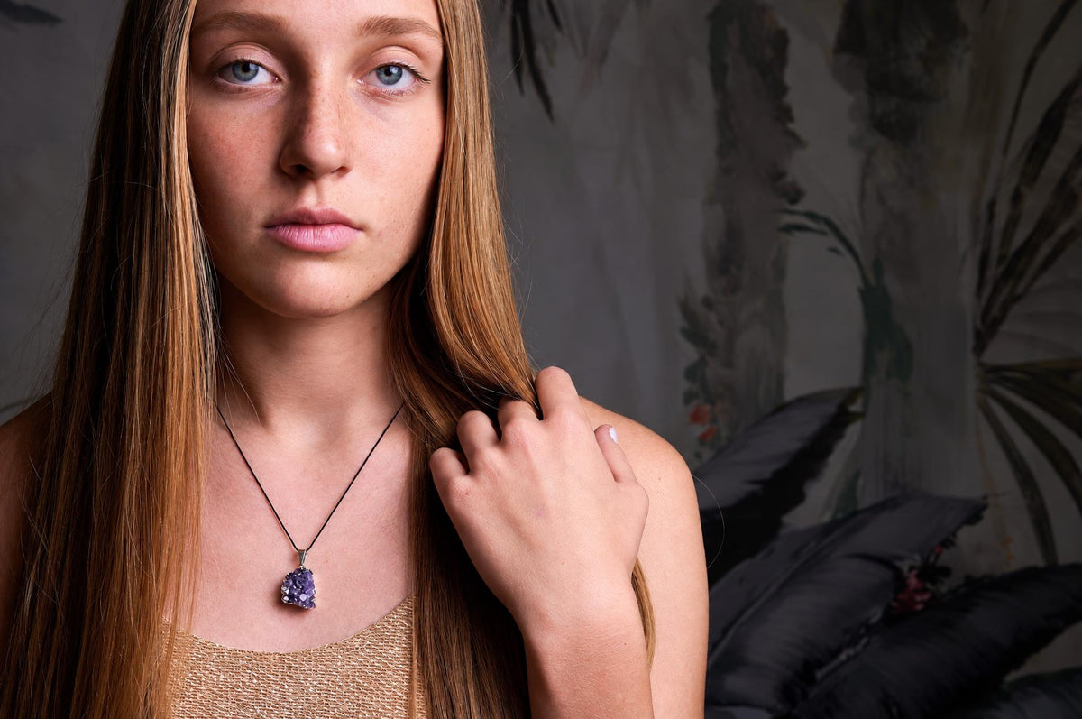 A woman wears a necklace with a purple amethyst pendant on a black cord against a dark surface.