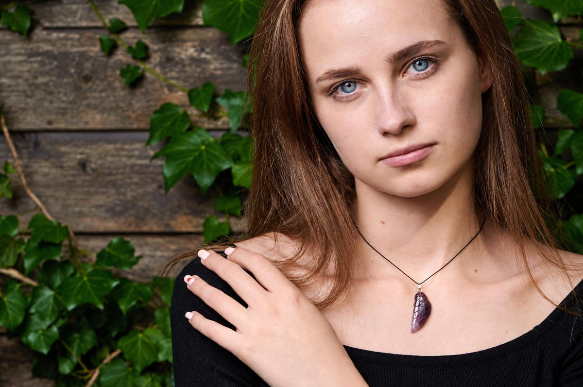 A woman wears a dark purple, wing-shaped pendant on a black cord against a wooden background with green ivy.
