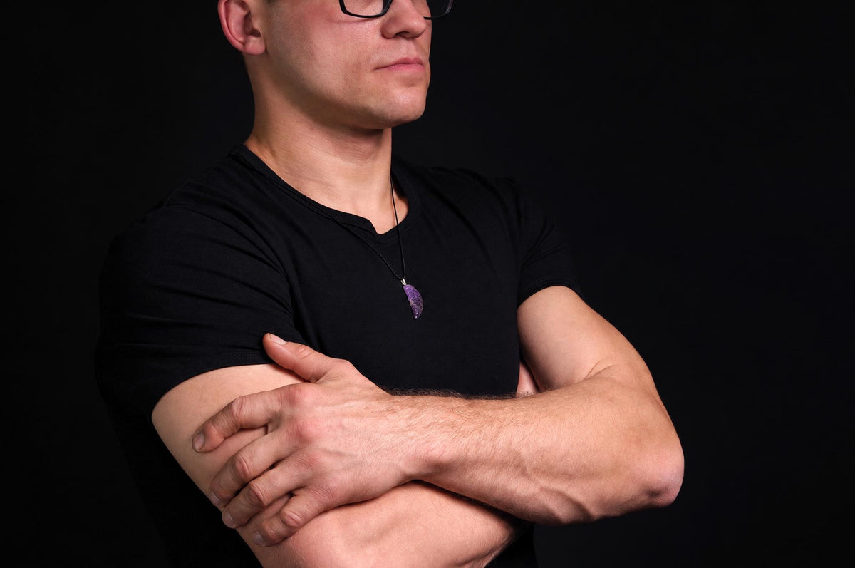 A man in a black shirt wears a necklace with a purple, wing-shaped pendant against a dark background.
