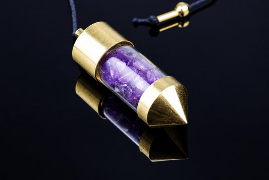 A brass pendulum with a clear tube containing purple amethyst stones, resting on a dark reflective surface.