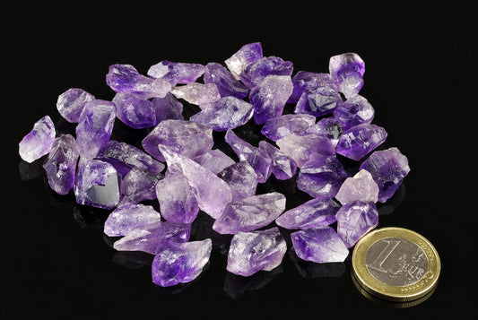 A pile of purple and clear amethyst rough crystals on a dark surface, next to a one-euro coin.
