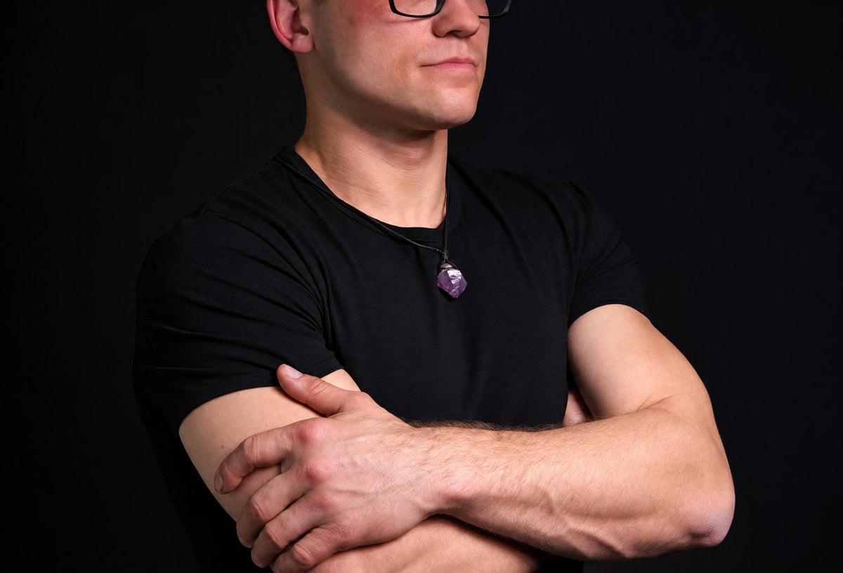 A man in a black t-shirt wears a necklace with a faceted purple amethyst pendant on a dark surface.