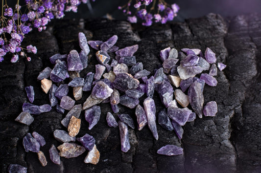 A pile of raw amethyst crystals in shades of purple and grey rests on a dark, textured surface with dried purple flowers nearby.