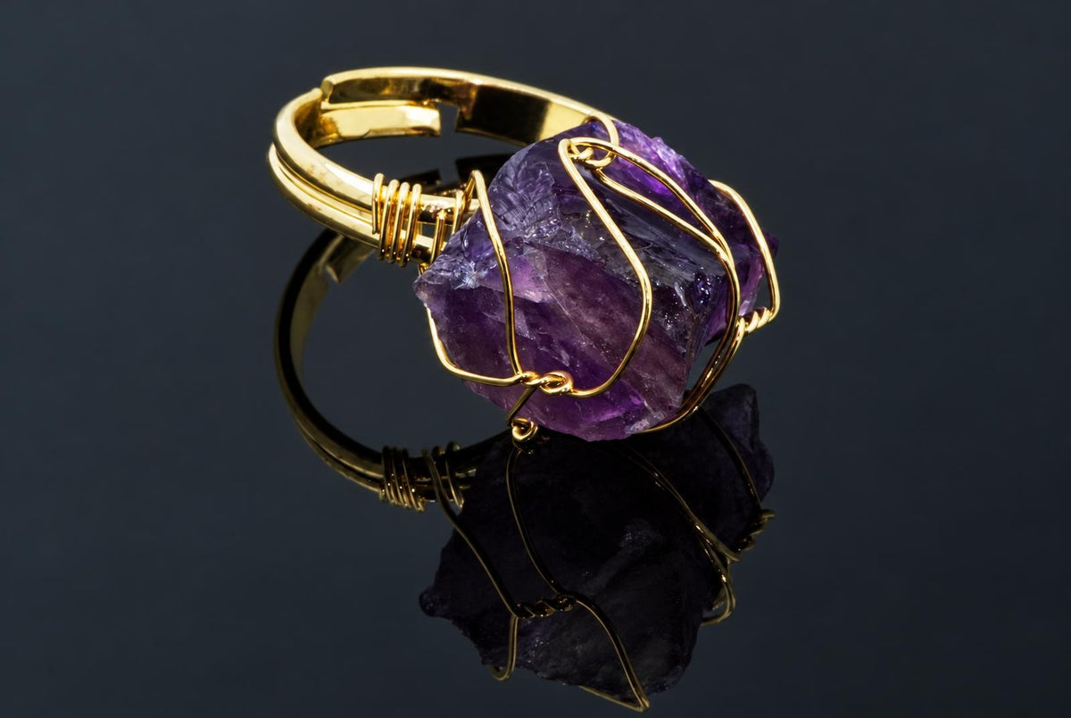 A gold-coloured ring with a rough purple crystal wrapped in thin wire rests on a dark reflective surface.
