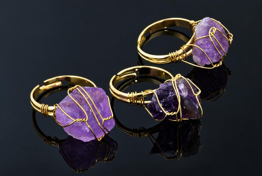 Gold-coloured rings hold rough purple crystals wrapped in thin wire on a dark reflective surface.