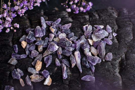 A pile of purple and grey amethyst crystals on a dark surface with dried purple flowers nearby.