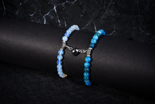 Two bracelets of pale blue and bright blue round beads are joined by a silver-tone heart charm on a black display.